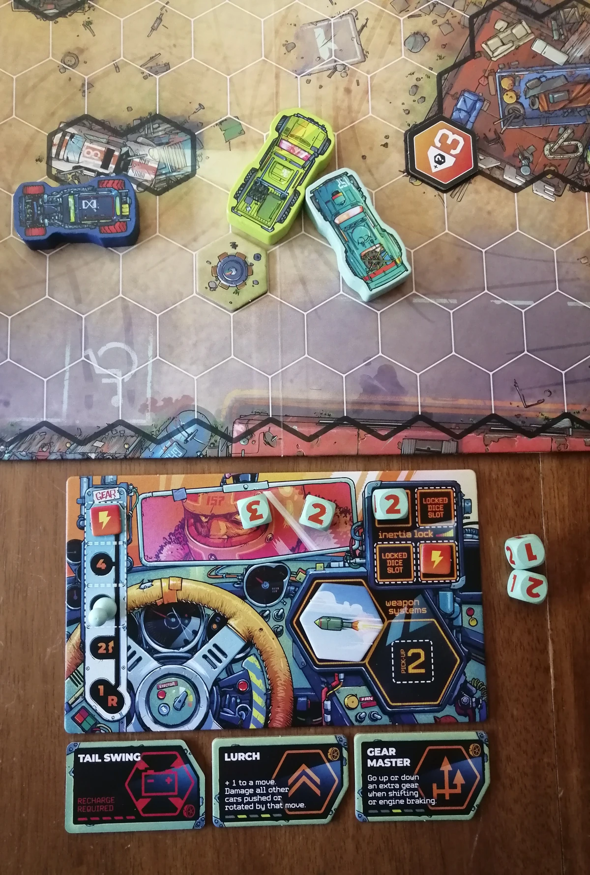 A photo of the board and a player set up next to it. The player set up includes a dashboard, which includes sections for placing dice, items and damage tokens. Dice are stored next to the dashboard, as well as on the large rearview mirror. Some dice are placed on locked dice slots, where they fit snuggly. The item tokens also fit snugly in their slot on the dashboard. Damage tokens are slightly smaller than their slots, but particularly in the locked dice slot section they need to be placed with accuracy. Below are three punchboard tokens for abilities which fit comfortably under the dashboard. One of the ability tokens is flipped to its recharge side.
