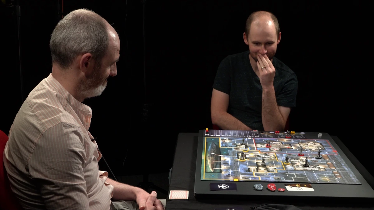 Two people playing Sniper Elite the board game. One is smiling, and one is looking at the board with a concerned smile and hand near their face.