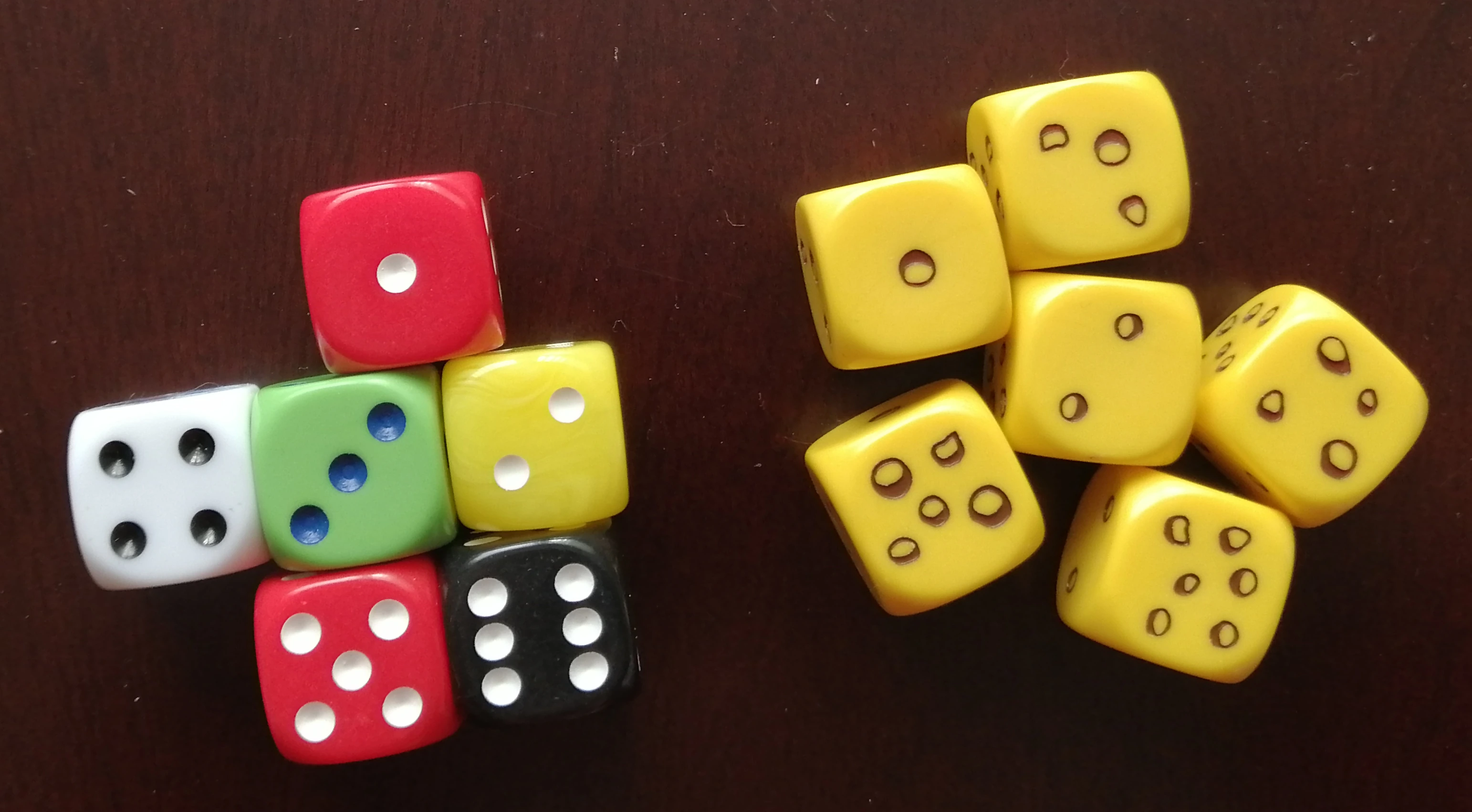 A photo of six-sided dice. On the left are a selection of solid coloured dice with pips from 1-6. On the right hand side are size of the mice dice - Tiny in the Tower themed dice. The mice dice are bright yellow with pips that resemble holes - giving the overall impression that the dice look like cheese.
